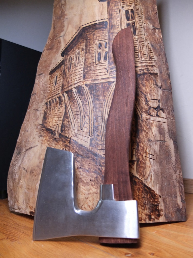 May include: A large, silver axe with a wooden handle. The axe is resting on a piece of wood with a burned image of a building.