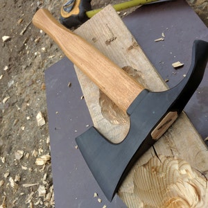 Op de afbeelding: Een zwarte en bruine handbijl met een houten steel. De bijl ligt op een stuk hout met houtsnippers eromheen.