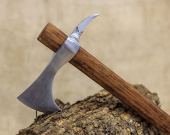 Peut inclure: Une hache de guerre en argent avec un manche en bois. La hache est posée sur un morceau de bois.