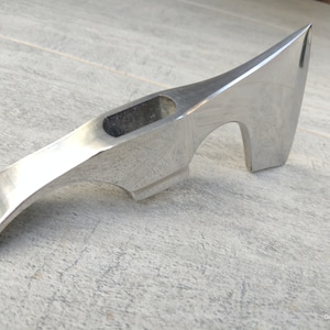 May include: A polished, silver-colored axe head with a curved handle and a sharp, pointed blade. The axe head features a rectangular opening near the top and a curved notch near the blade. The tool is designed for chopping and splitting.