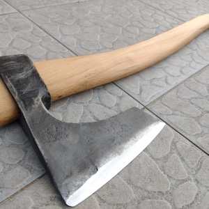 Op de afbeelding: Een handgesmede bijl met een lichtgekleurde houten steel. De bijlkop is gemaakt van donker metaal met een scherp, zilverkleurig blad. De steel buigt zachtjes.