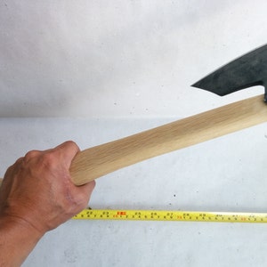 Peut inclure: Une hache à manche en bois avec une lame en métal noir. La hache est tenue par une main, et un ruban à mesurer jaune est visible au premier plan.