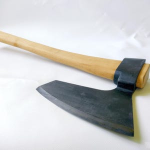 Puede incluir: Hacha con mango de madera marrón claro y hoja de metal oscuro. La cabeza del hacha está unida al mango con un collar de metal negro. El hacha está diseñada para cortar madera.