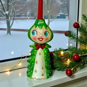May include: A whimsical ceramic Christmas tree candle holder with a red candle. The holder is green with white polka dots and a smiling face with blue eyes. A red bow adorns the neck. The background shows a snowy winter scene.