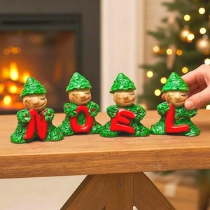 May include: Four ceramic elf figurines spelling "NOEL" in red letters. The elves have green textured outfits and hats, with smiling faces. The figurines are on a wooden table, with a fireplace and Christmas tree in the background.