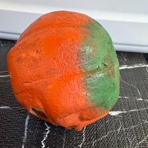 May include: A textured, rounded object with a vibrant color scheme. The top half is painted bright orange, transitioning to green on the right side. The object sits on a black surface with white veining.