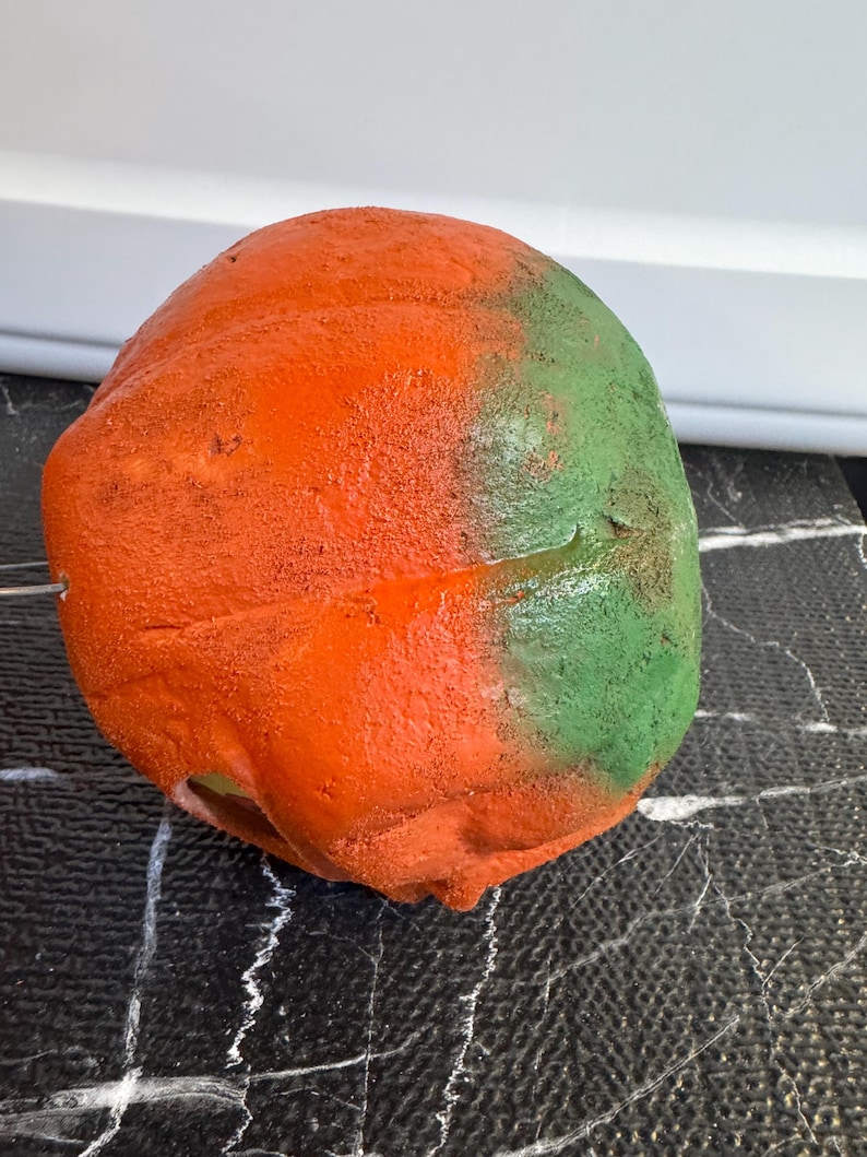 May include: A textured, rounded object with a vibrant color scheme. The top half is painted bright orange, transitioning to green on the right side. The object sits on a black surface with white veining.