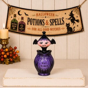 May include: A purple ceramic potion bottle with a bat-shaped stopper featuring a pale face and black wings. The bottle has a textured, bulbous body with the text BAT WINGS printed in black on the front. The design includes a pointed lid.