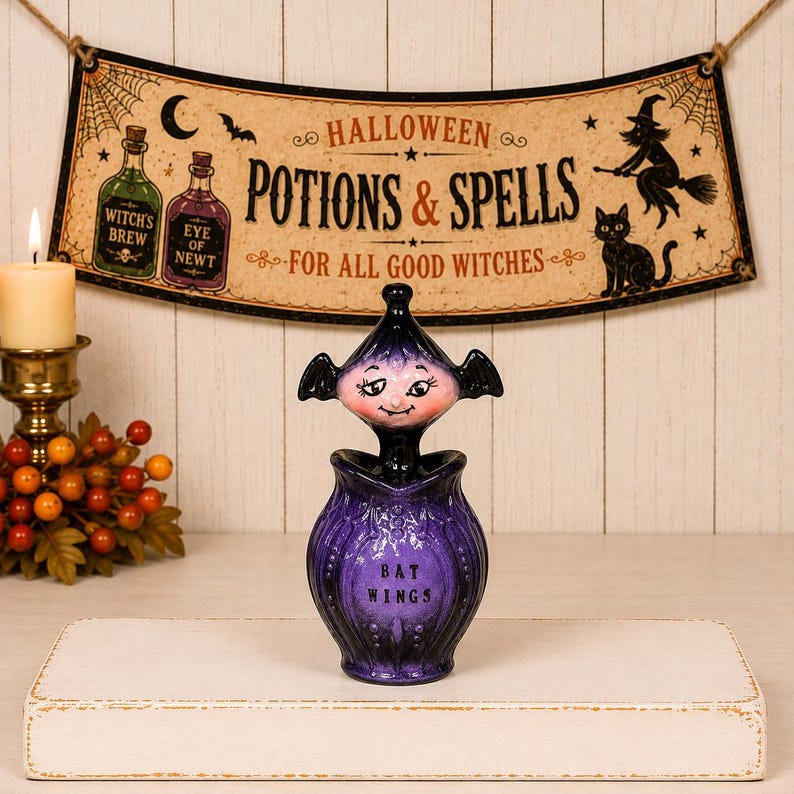 May include: A purple ceramic potion bottle with a bat-shaped stopper featuring a pale face and black wings. The bottle has a textured, bulbous body with the text BAT WINGS printed in black on the front. The design includes a pointed lid.