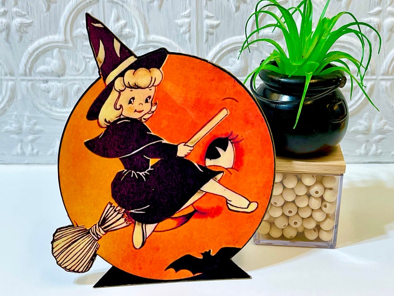 May include: A vintage Halloween decoration featuring a witch riding a broomstick on a large orange moon. The witch is wearing a black dress and a pointed hat. The moon has a large eye and a bat flying in front of it.