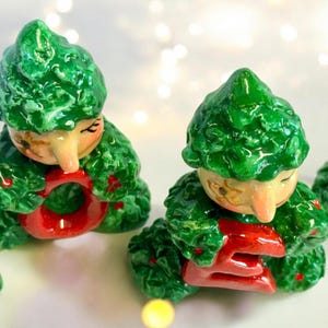 May include: Ceramic figurines of seated elves with green leafy outfits and red accents. The elves have tan faces and are holding red letters. The figurines are likely decorative items.