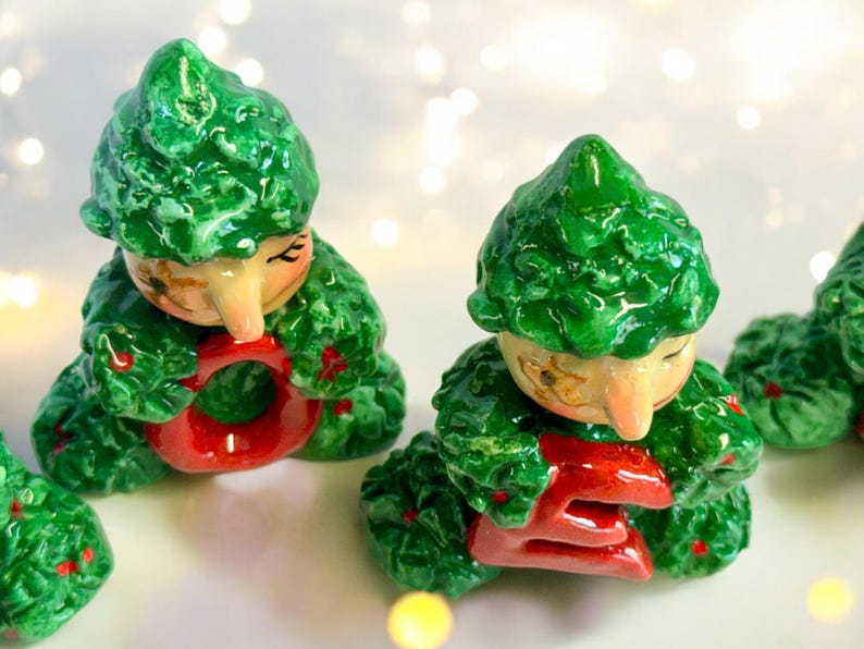 May include: Ceramic figurines of seated elves with green leafy outfits and red accents. The elves have tan faces and are holding red letters. The figurines are likely decorative items.