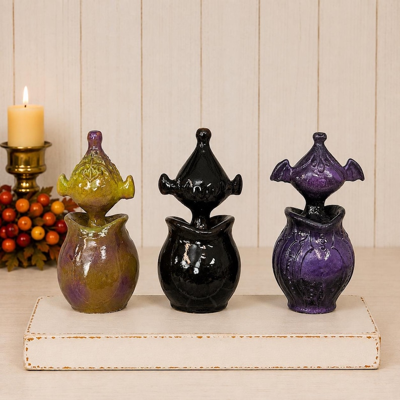 May include: Three ceramic decorative vessels with lids, each featuring a distinct shape and finish. One is a mottled olive green and purple, one is a solid glossy black, and one is a deep purple with a textured surface and wing-like accents.