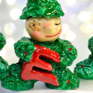 May include: A ceramic figurine of a smiling character with a green leafy outfit and hat, holding a large red letter "E". The face is painted with rosy cheeks, closed eyes, and a red smile. A star is painted on the face.