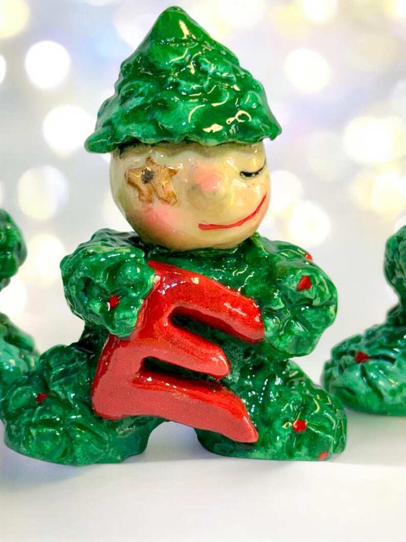 May include: A ceramic figurine of a smiling character with a green leafy outfit and hat, holding a large red letter "E". The face is painted with rosy cheeks, closed eyes, and a red smile. A star is painted on the face.