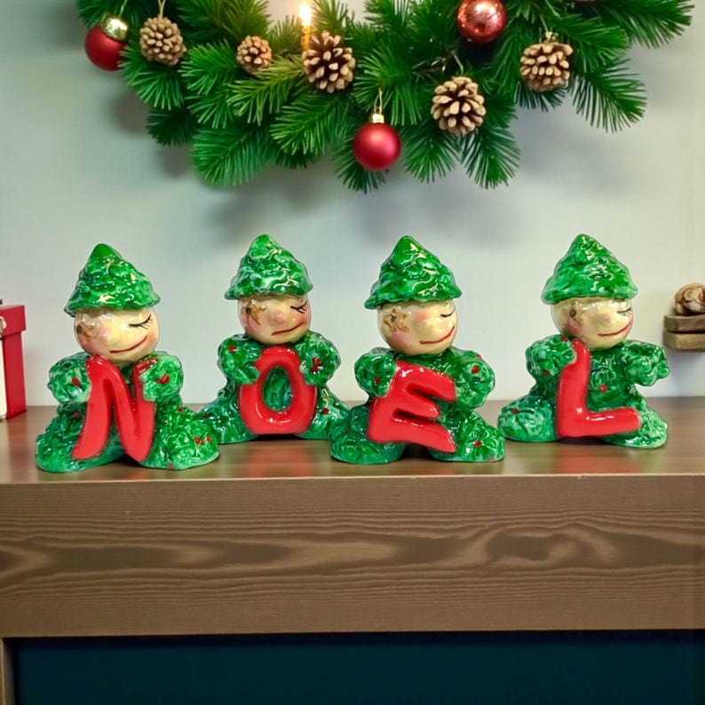May include: Four ceramic figures spelling "NOEL" in red letters. The figures are dressed in green, with green hats and smiling faces. A Christmas wreath with baubles and pinecones is in the background.