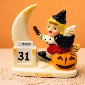 May include: A ceramic Halloween calendar figurine featuring a blonde character in a black witch hat and cape riding a broomstick atop a smiling orange jack-o'-lantern. A white crescent moon stands behind, with a block displaying October 31.