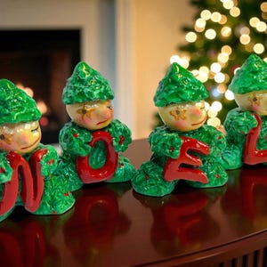 May include: Four Christmas figurines spelling "NOEL". Each figurine has a green leafy body and hat, with a painted face. The letters are red. The figurines are on a dark brown surface, with a fireplace and Christmas tree in the background.