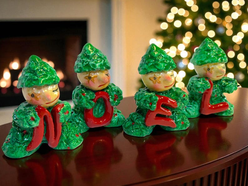 May include: Four Christmas figurines spelling "NOEL". Each figurine has a green leafy body and hat, with a painted face. The letters are red. The figurines are on a dark brown surface, with a fireplace and Christmas tree in the background.