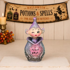 May include: A ceramic figurine featuring a winking pixie with purple hair emerging from a rounded vessel. The vessel is decorated with raised star patterns and the text PIXIE DUST in black lettering against a pink background with purple accents.
