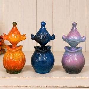 May include: Three ceramic jars with lids, each featuring a rounded base and a pointed, finial-style top. The first jar is orange, the second is deep blue, and the third is a gradient of purple and blue. Each piece has a textured, glossy finish.