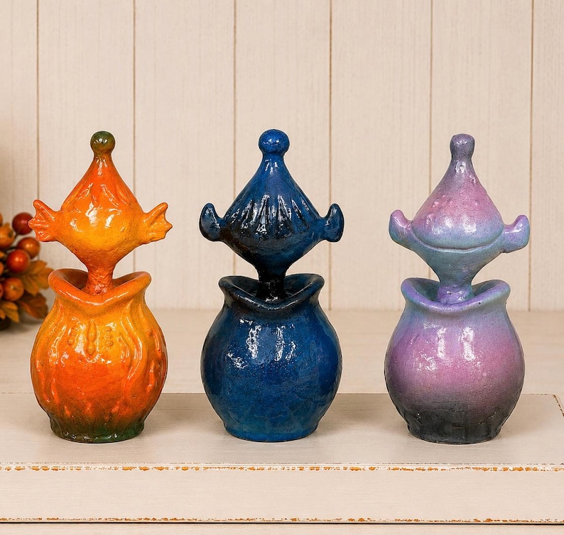 May include: Three ceramic jars with lids, each featuring a rounded base and a pointed, finial-style top. The first jar is orange, the second is deep blue, and the third is a gradient of purple and blue. Each piece has a textured, glossy finish.