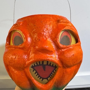 May include: A vintage orange Halloween pumpkin pail with a grinning face, large eyes, and a jagged mouth. The pail has a wire handle and a green base. The pumpkin's face is made of paper and has a textured appearance.