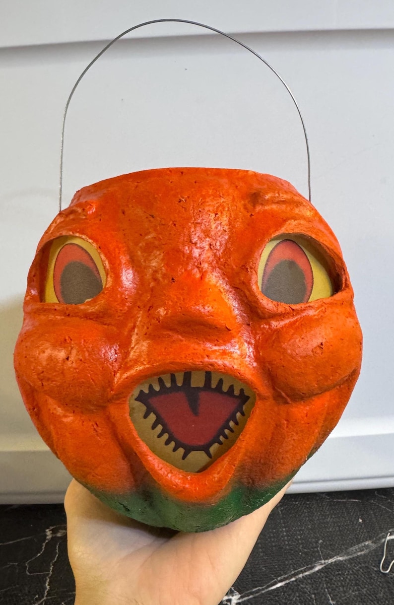 May include: A vintage orange Halloween pumpkin pail with a grinning face, large eyes, and a jagged mouth. The pail has a wire handle and a green base. The pumpkin's face is made of paper and has a textured appearance.