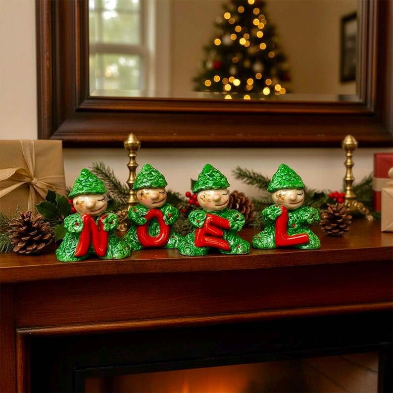 May include: Four ceramic elf figurines spelling "NOEL" in red letters. The elves wear green hats and outfits with gold faces. The figurines are displayed on a wooden mantelpiece with a Christmas tree in the background.