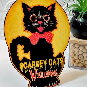 May include: A black cat with big eyes and a red polka dot bow tie is on a yellow moon with the text "Scardey Cats Welcome" dripping in red and orange.
