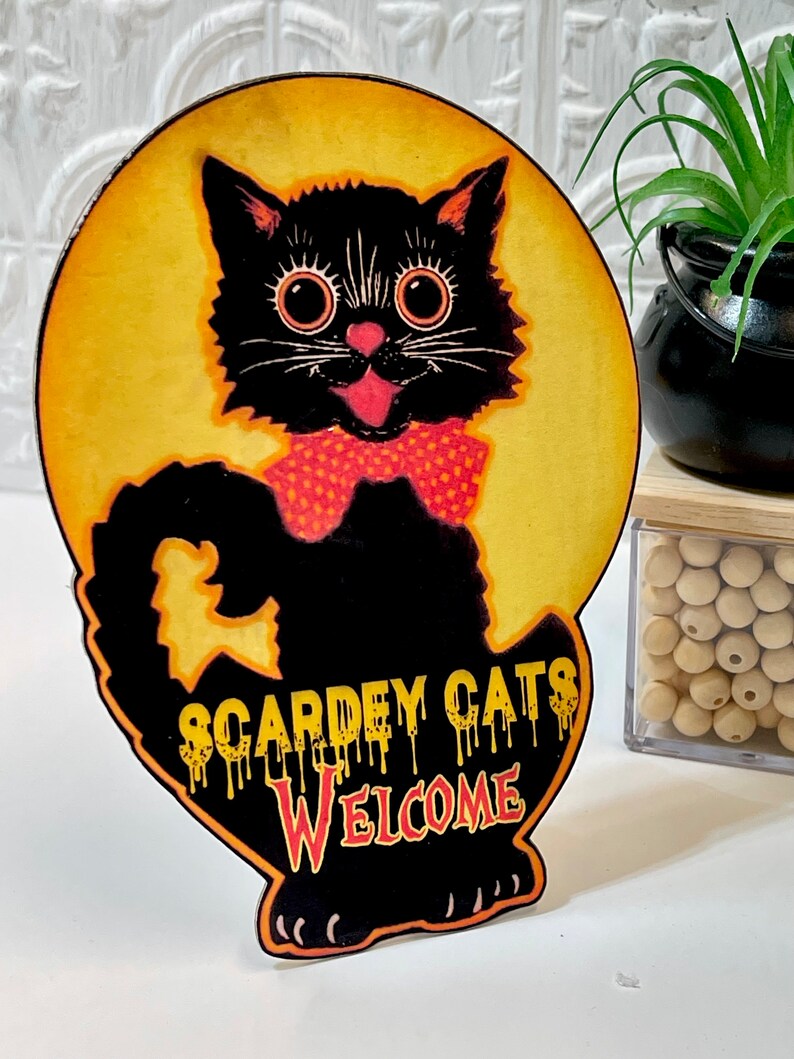 May include: A black cat with big eyes and a red polka dot bow tie is on a yellow moon with the text "Scardey Cats Welcome" dripping in red and orange.