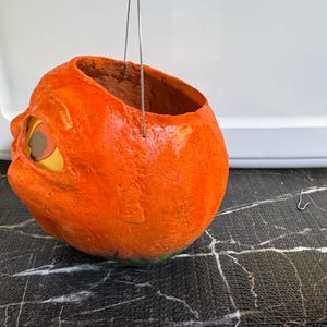 May include: A handmade, orange pumpkin-shaped container with a wide opening and a wire handle. The pumpkin has a textured surface, a carved eye, and a green base. Perfect for Halloween decorations or as a unique storage item.