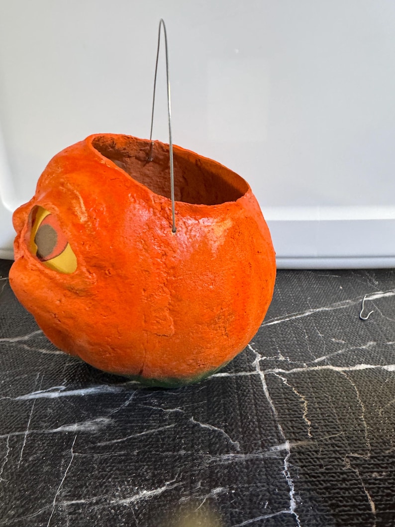 May include: A handmade, orange pumpkin-shaped container with a wide opening and a wire handle. The pumpkin has a textured surface, a carved eye, and a green base. Perfect for Halloween decorations or as a unique storage item.