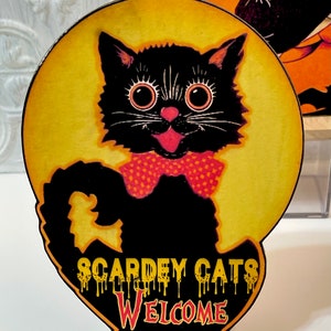 May include: A black cat with big eyes and a red bow tie is silhouetted against a yellow moon. The text "Scardey Cats Welcome" is written in dripping yellow letters on the cat's body.