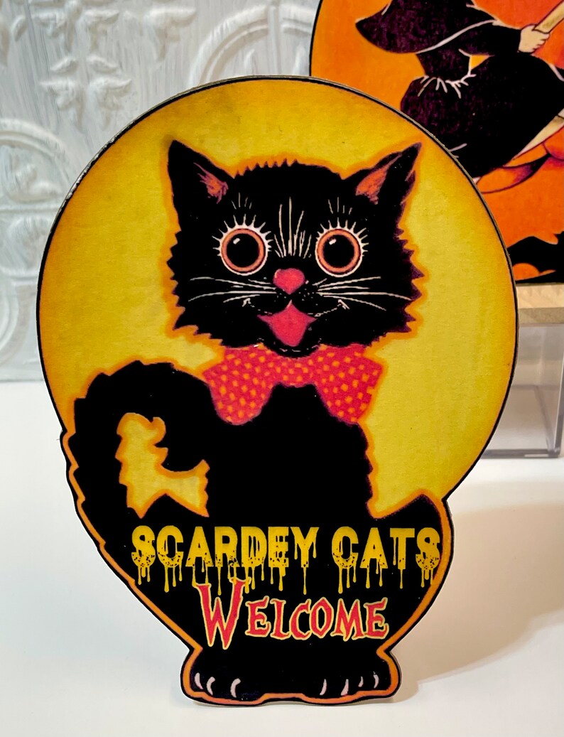 May include: A black cat with big eyes and a red bow tie is silhouetted against a yellow moon. The text "Scardey Cats Welcome" is written in dripping yellow letters on the cat's body.