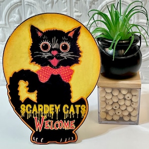 May include: A wooden Halloween sign with a black cat illustration and the text "Scardey Cats Welcome". The cat is wearing a red polka dot bow tie and has large, round eyes. The sign is painted with a yellow background and dripping orange paint.