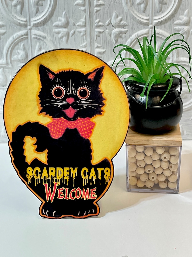 May include: A wooden Halloween sign with a black cat illustration and the text "Scardey Cats Welcome". The cat is wearing a red polka dot bow tie and has large, round eyes. The sign is painted with a yellow background and dripping orange paint.