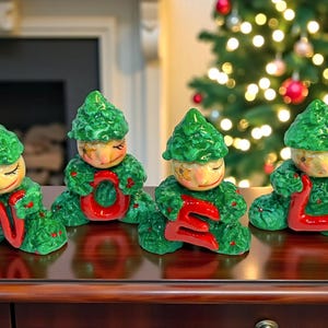 May include: Four ceramic Christmas figurines spelling "NOEL". Each figurine is a green-clad elf holding a red letter. The elves have painted faces and are posed on a dark wooden surface, with a Christmas tree in the background.