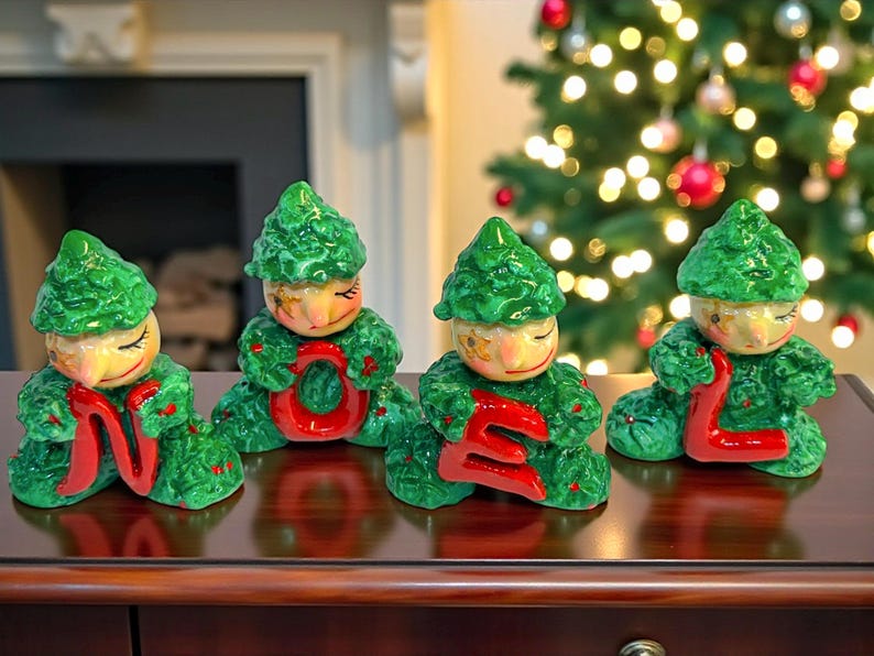May include: Four ceramic Christmas figurines spelling "NOEL". Each figurine is a green-clad elf holding a red letter. The elves have painted faces and are posed on a dark wooden surface, with a Christmas tree in the background.