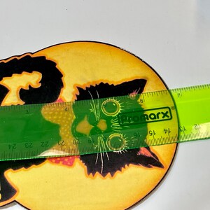 May include: A round, yellow and orange Halloween-themed coaster with a black cat illustration and the text "SCARE-EM RATS". The coaster is being measured with a green ruler.