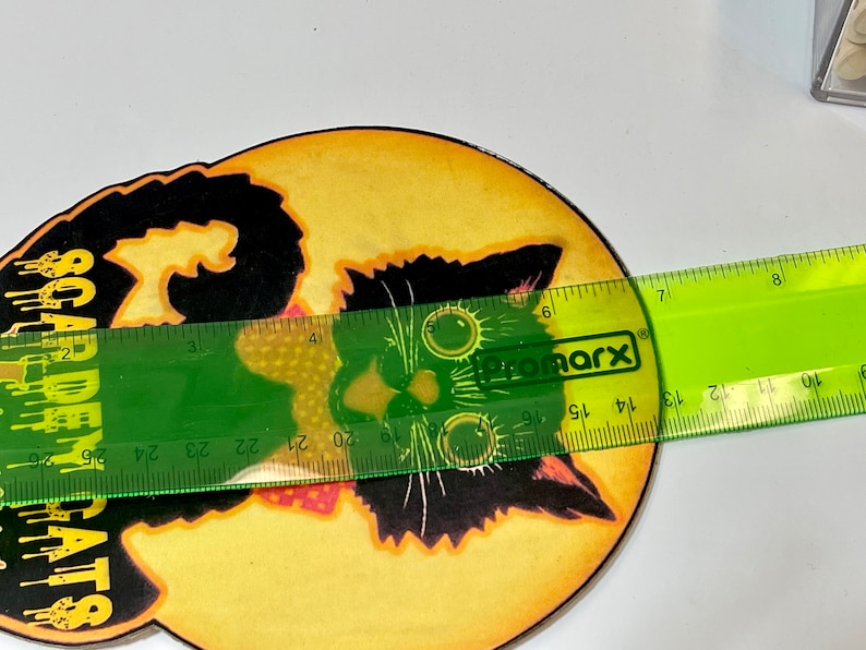 May include: A round, yellow and orange Halloween-themed coaster with a black cat illustration and the text "SCARE-EM RATS". The coaster is being measured with a green ruler.