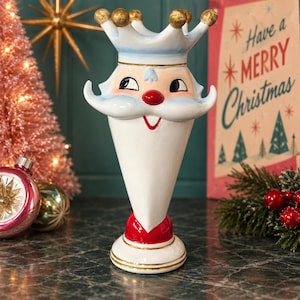 May include: A ceramic vase shaped like a stylized Santa Claus head with a crown. The figure features a white beard, a red nose, a red mouth, and gold-painted accents on the crown points and base. The surface is glossy white with blue shading.