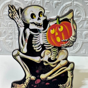 May include: A vintage Halloween decoration featuring a skeleton holding a jack-o'-lantern. The skeleton is sitting on a black background with red mushrooms.
