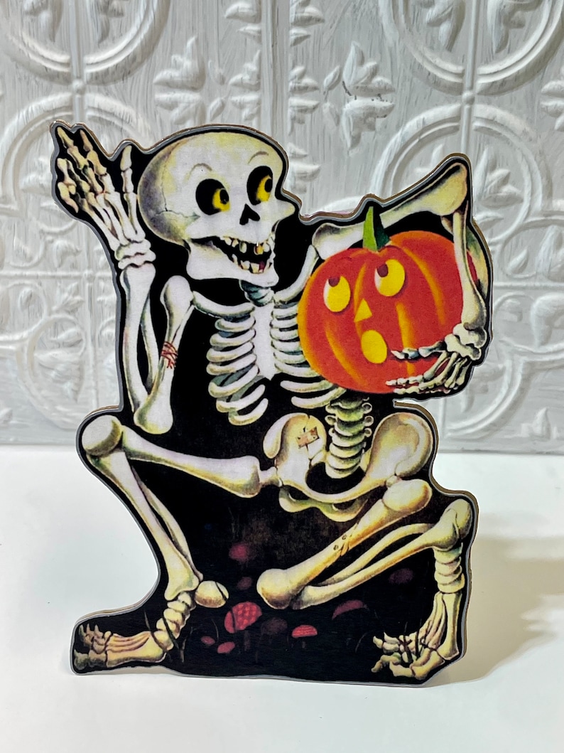 May include: A vintage Halloween decoration featuring a skeleton holding a jack-o'-lantern. The skeleton is sitting on a black background with red mushrooms.