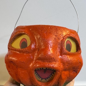 May include: Vintage orange Halloween pumpkin pail with a grinning face, yellow eyes, and a wire handle. The pail has a textured surface and a green base, perfect for trick-or-treating.