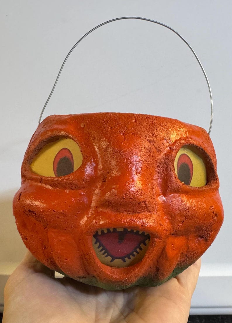 May include: Vintage orange Halloween pumpkin pail with a grinning face, yellow eyes, and a wire handle. The pail has a textured surface and a green base, perfect for trick-or-treating.