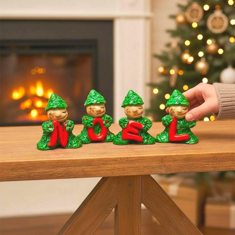 May include: Four ceramic elf figurines spelling "NOEL" in red letters. The elves have green textured outfits and hats, with smiling faces. The figurines are on a wooden table, with a fireplace and Christmas tree in the background.