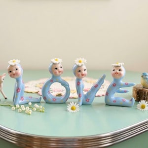 May include: A light blue ceramic sculpture spells out the word "LOVE" with each letter shaped like a doll figure. The dolls wear daisy hats and have pink floral accents. A small deer and bird figurine are also present.