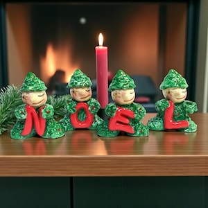 May include: A Christmas-themed display featuring four ceramic figures spelling "NOEL" in red letters. The figures are dressed in green outfits and hats, with smiling faces. A red candle is lit in the background, with a fireplace out of focus.