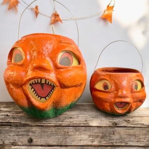 May include: Two orange Halloween pumpkin buckets with smiling faces and wire handles. The larger pumpkin has a green base and a wide open mouth with teeth. The smaller pumpkin has a similar design. Both are on a weathered wooden surface.
